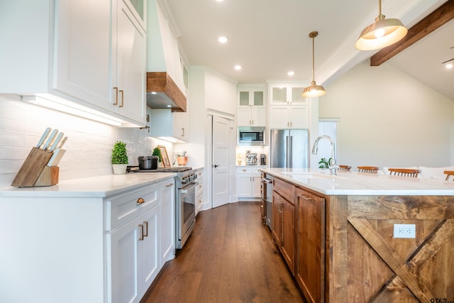 kitchen featuring appliances with stainless steel finishes, custom range hood, pendant lighting, a center island with sink, and white cabinetry