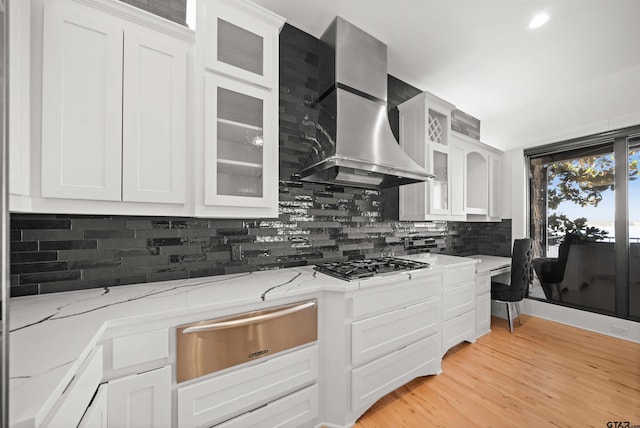 kitchen with white cabinetry, wall chimney range hood, light stone countertops, a warming drawer, and glass insert cabinets