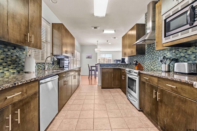 kitchen with light tile patterned floors, a peninsula, dark stone counters, stainless steel appliances, and wall chimney range hood