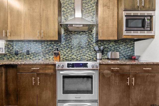 kitchen with light stone counters, stainless steel appliances, dark brown cabinets, wall chimney exhaust hood, and backsplash