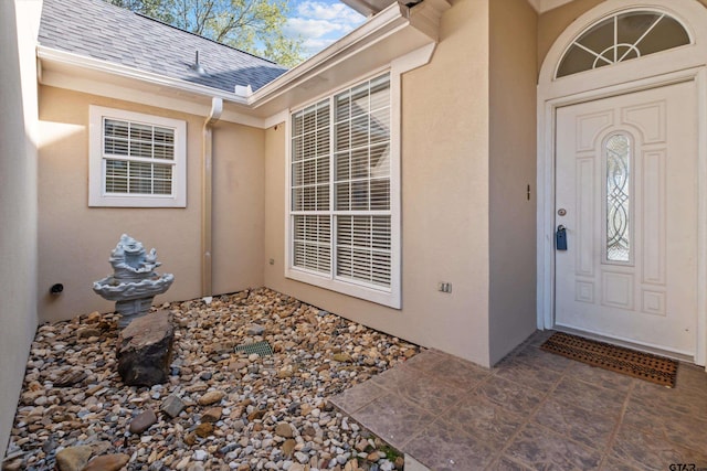 view of exterior entry with stucco siding and a shingled roof