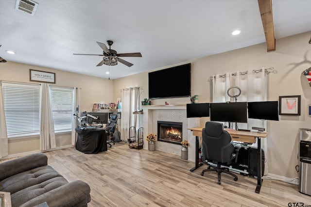 home office featuring beamed ceiling, a tiled fireplace, ceiling fan, and light hardwood / wood-style flooring