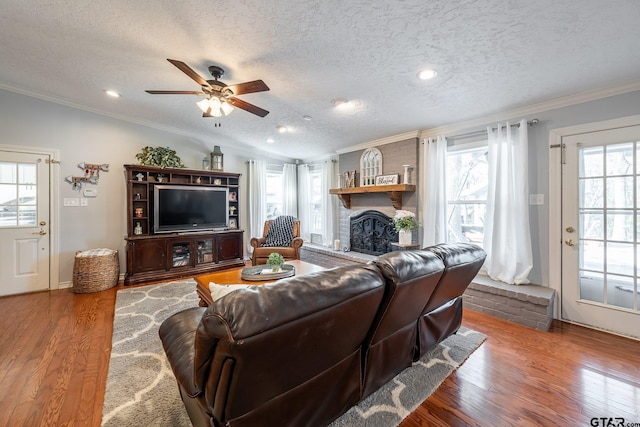 living room with wood-type flooring, a brick fireplace, a textured ceiling, ornamental molding, and ceiling fan