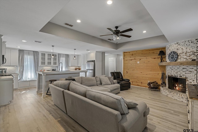 living room featuring ceiling fan, sink, a tray ceiling, a fireplace, and light hardwood / wood-style floors