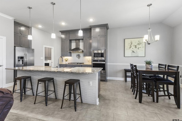 kitchen with appliances with stainless steel finishes, a kitchen bar, wall chimney range hood, hanging light fixtures, and a center island with sink