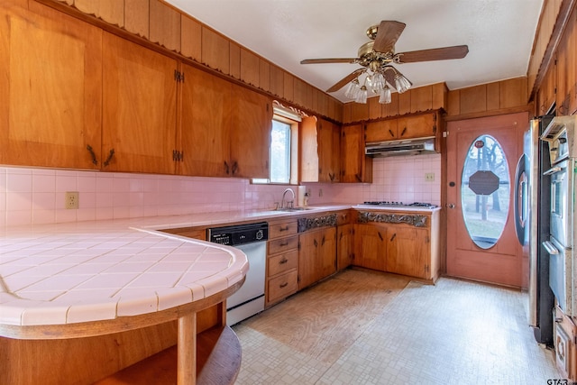 kitchen with tile counters, brown cabinetry, a sink, under cabinet range hood, and dishwashing machine