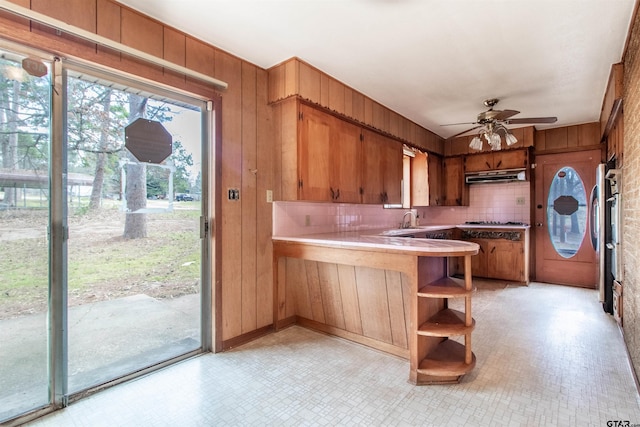 kitchen featuring brown cabinetry, a sink, wood walls, a peninsula, and under cabinet range hood