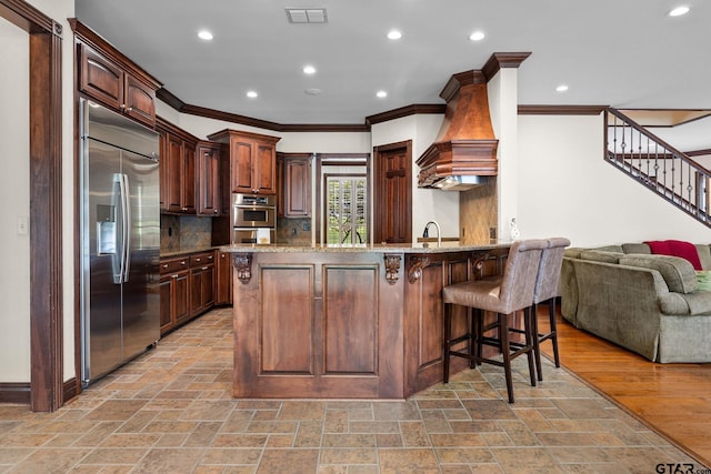 kitchen featuring a breakfast bar area, appliances with stainless steel finishes, light stone counters, custom exhaust hood, and kitchen peninsula