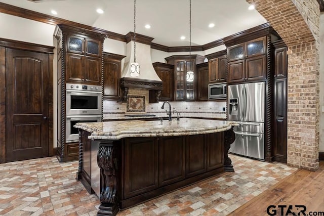 kitchen featuring appliances with stainless steel finishes, custom range hood, decorative light fixtures, an island with sink, and dark brown cabinets