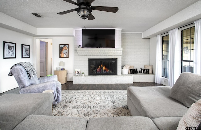 living room with wood finished floors, a ceiling fan, a brick fireplace, a textured ceiling, and visible vents