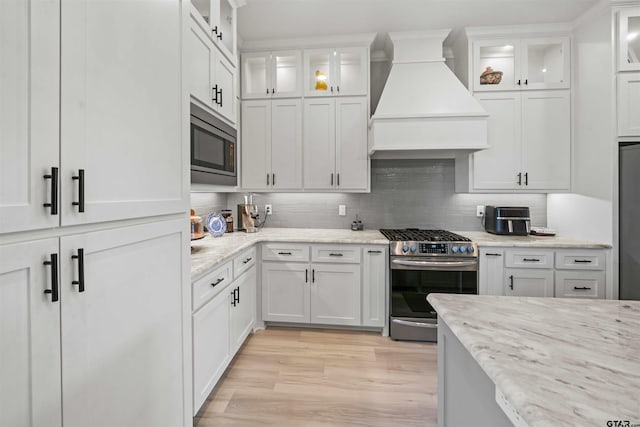 kitchen with stainless steel appliances, light wood-type flooring, custom range hood, decorative backsplash, and white cabinets