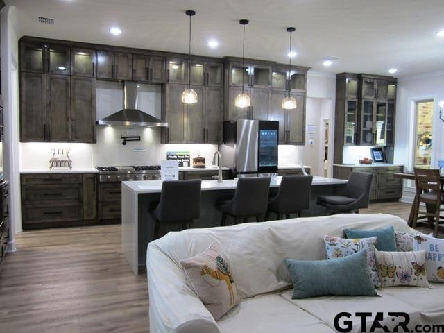 kitchen with a kitchen island with sink, wall chimney range hood, appliances with stainless steel finishes, pendant lighting, and light hardwood / wood-style flooring