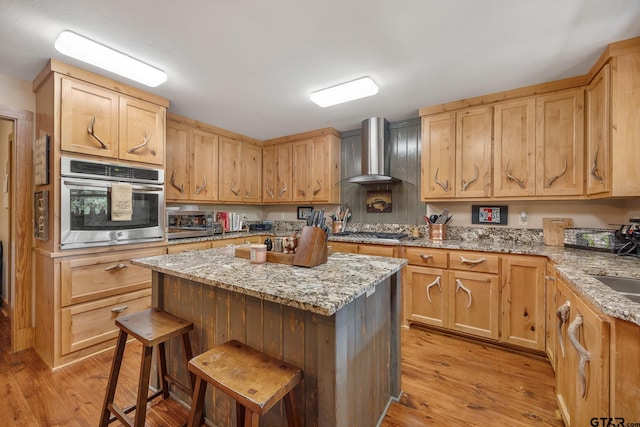 kitchen featuring wall chimney range hood, appliances with stainless steel finishes, light stone countertops, a breakfast bar area, and light hardwood / wood-style flooring