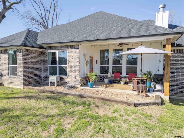 back of property featuring a shingled roof, a ceiling fan, a chimney, a yard, and brick siding