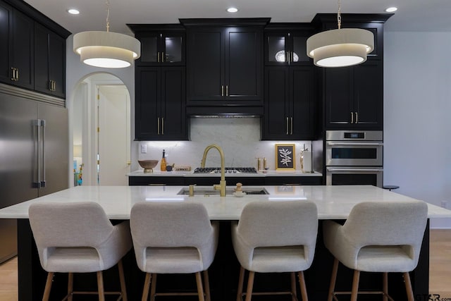 kitchen featuring glass insert cabinets, stainless steel appliances, a center island with sink, and dark cabinets