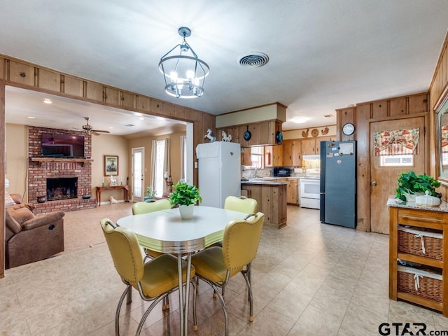 dining space featuring ceiling fan with notable chandelier, wooden walls, sink, and a brick fireplace