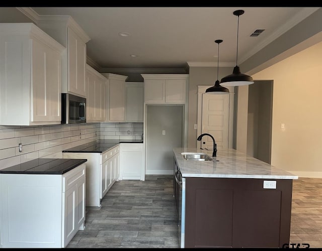 kitchen featuring stainless steel microwave, hanging light fixtures, white cabinetry, a sink, and an island with sink