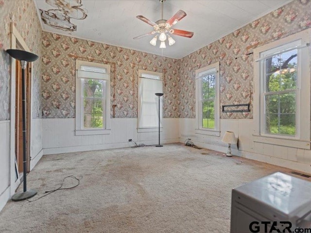 carpeted empty room with ceiling fan, ornamental molding, wainscoting, and a wealth of natural light