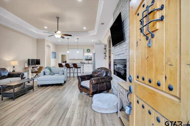 living room with crown molding, a brick fireplace, light hardwood / wood-style flooring, a tray ceiling, and ceiling fan