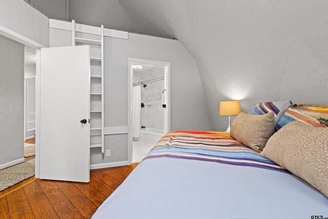 bedroom with a textured wall, lofted ceiling, baseboards, connected bathroom, and hardwood / wood-style flooring
