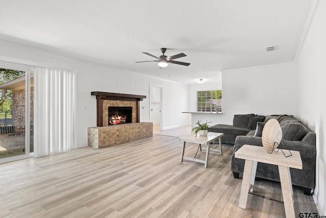living room featuring crown molding, light wood-style floors, a ceiling fan, visible vents, and a brick fireplace