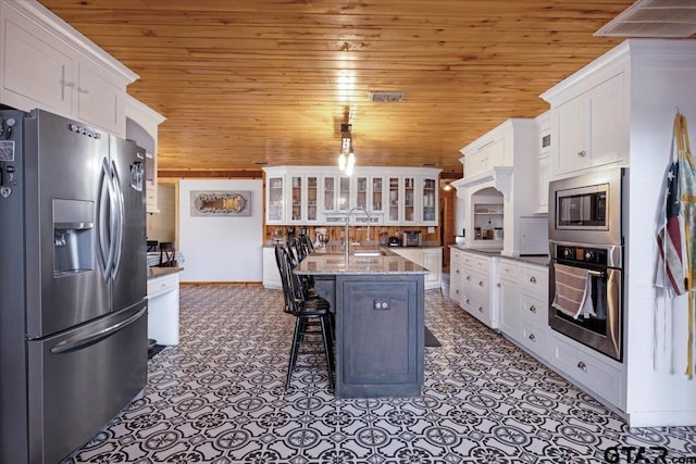 kitchen featuring appliances with stainless steel finishes, white cabinetry, a kitchen breakfast bar, a center island with sink, and wooden ceiling