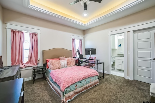 bedroom with ensuite bathroom, a ceiling fan, baseboards, a tray ceiling, and dark carpet