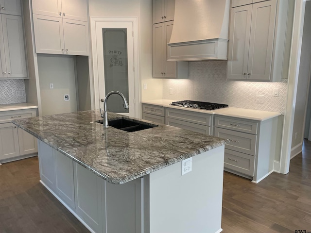 kitchen with custom range hood, stainless steel gas cooktop, sink, dark hardwood / wood-style floors, and an island with sink