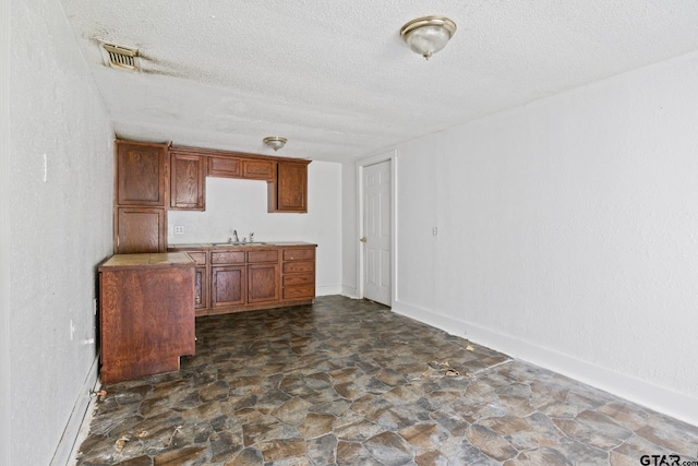 kitchen featuring a sink, a textured ceiling, brown cabinetry, stone finish floor, and visible vents