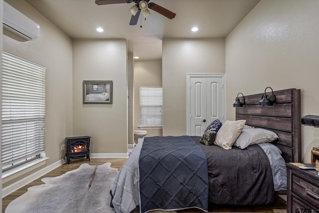 bedroom featuring ensuite bath, a wall mounted air conditioner, ceiling fan, light hardwood / wood-style flooring, and a wood stove