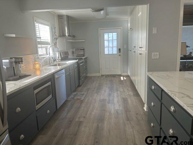 kitchen featuring light stone counters, stainless steel appliances, wood finished floors, a sink, and wall chimney exhaust hood