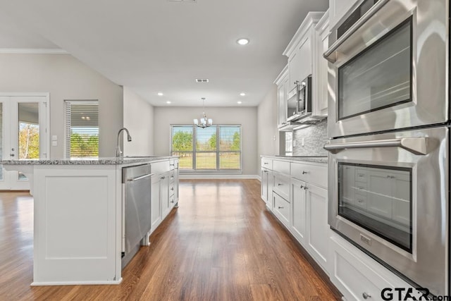 kitchen featuring appliances with stainless steel finishes, light stone counters, a center island with sink, white cabinets, and a chandelier