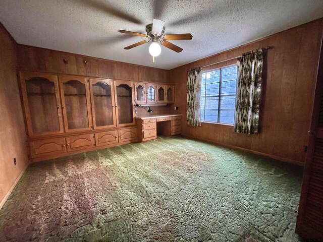 interior space with a textured ceiling, dark colored carpet, built in desk, and ceiling fan