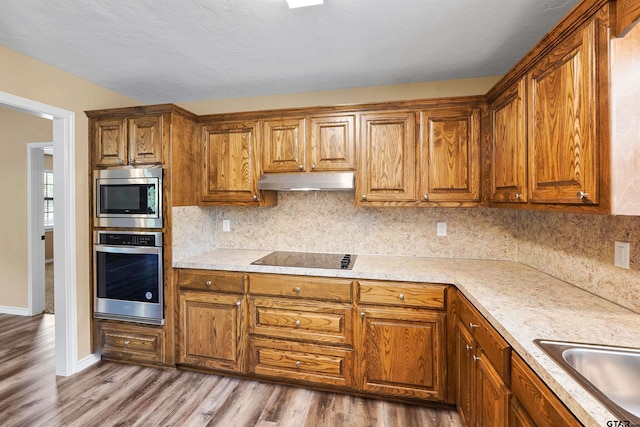 kitchen with appliances with stainless steel finishes, brown cabinets, wood finished floors, a sink, and under cabinet range hood