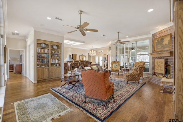 living room with ornamental molding, wood-type flooring, ceiling fan with notable chandelier, and a brick fireplace