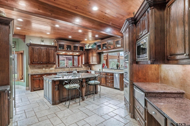 kitchen with a kitchen bar, an island with sink, stainless steel microwave, stone counters, and dark brown cabinetry