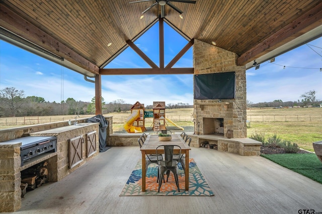 view of patio with a ceiling fan, fence, a playground, an outdoor stone fireplace, and an outdoor kitchen