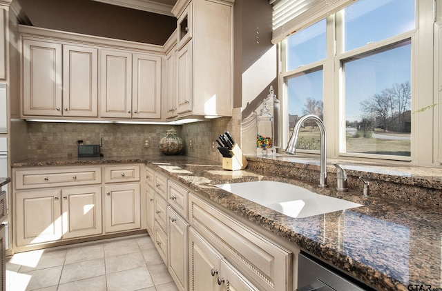 kitchen featuring cream cabinets, light tile patterned flooring, a sink, decorative backsplash, and dark stone counters