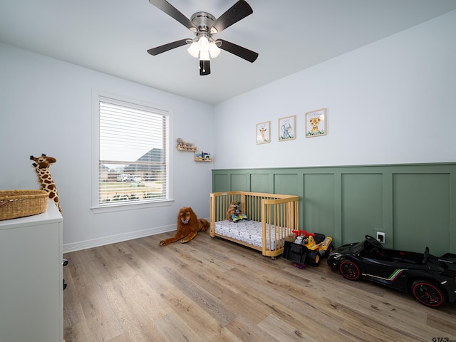 bedroom featuring a crib, ceiling fan, a wainscoted wall, light wood-style floors, and a decorative wall