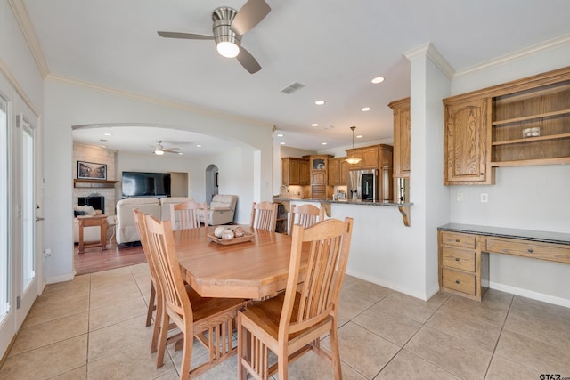 tiled dining space featuring ceiling fan, built in desk, a stone fireplace, and crown molding