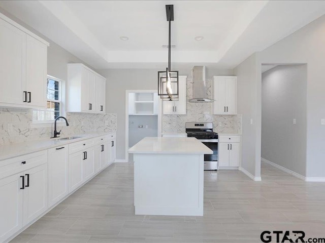 kitchen featuring a tray ceiling, a center island, stainless steel gas stove, a sink, and wall chimney range hood