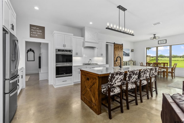 kitchen with white cabinetry, stainless steel appliances, a barn door, an island with sink, and concrete floors