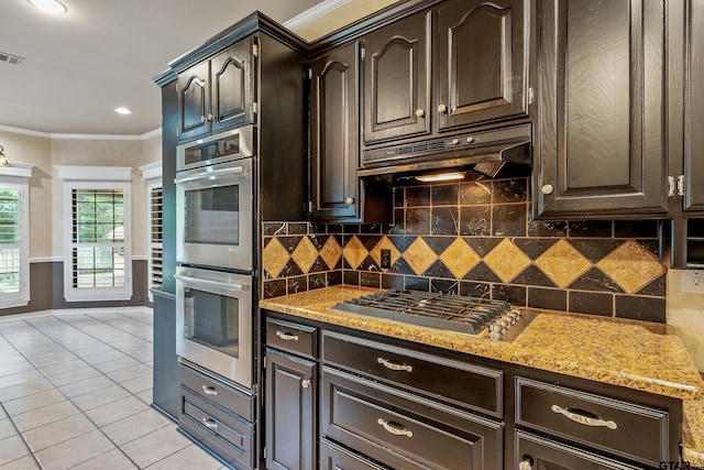 kitchen with light tile patterned flooring, ornamental molding, under cabinet range hood, appliances with stainless steel finishes, and backsplash