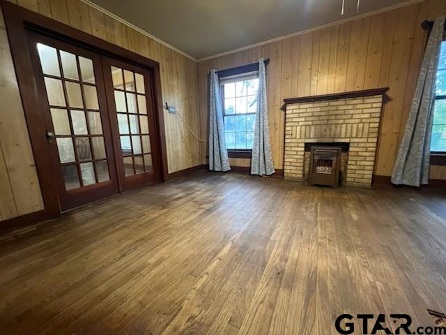 unfurnished living room with ornamental molding, a fireplace, wooden walls, wood finished floors, and french doors
