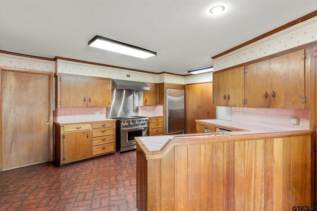 kitchen featuring wall chimney exhaust hood, premium appliances, decorative backsplash, brick floor, and tile countertops