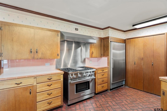 kitchen featuring tile counters, premium appliances, brick floor, decorative backsplash, and wall chimney exhaust hood