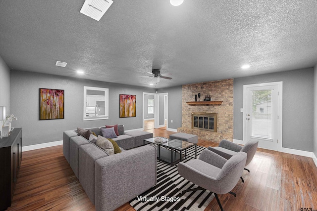 living room with a brick fireplace, dark wood-type flooring, a textured ceiling, and ceiling fan