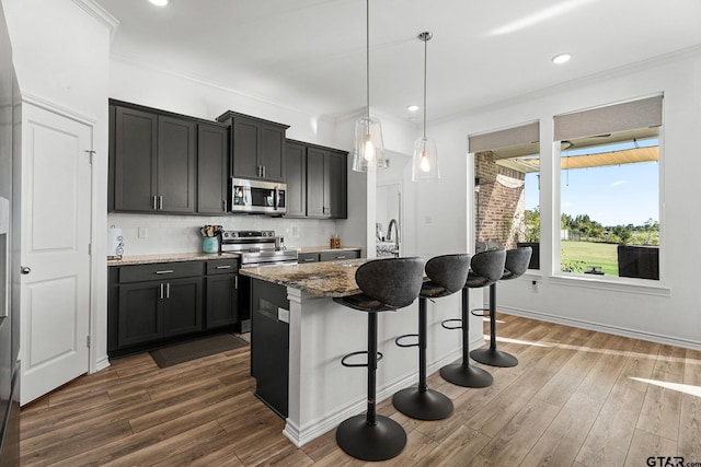 kitchen with dark stone countertops, a kitchen island with sink, stainless steel appliances, pendant lighting, and dark hardwood / wood-style floors