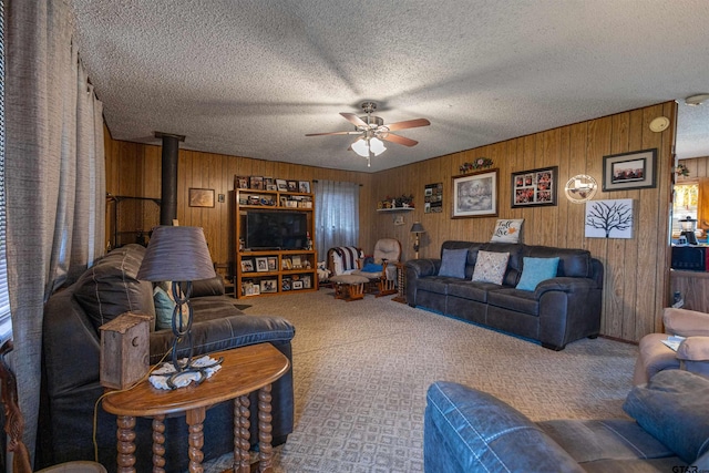 living room featuring ceiling fan, a textured ceiling, carpet flooring, a wood stove, and wooden walls