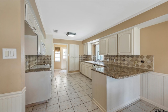 kitchen with kitchen peninsula, ornamental molding, dark stone countertops, light tile patterned floors, and backsplash